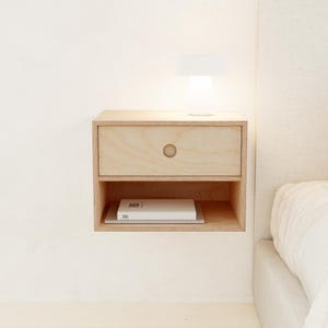 May include: A light-colored, floating nightstand with a drawer and open shelf. A small, white lamp sits on top of the nightstand. A book is visible on the shelf. The nightstand is mounted on a white wall next to a bed.
