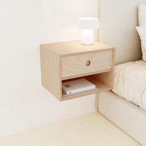 May include: A light-colored, floating nightstand made of wood. It features a drawer with a circular handle and an open shelf below. A small white lamp sits on top, and a book rests on the shelf. The nightstand is mounted on a white wall.