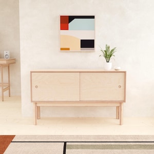 May include: A light wood-toned cabinet with sliding doors and a minimalist design. The cabinet sits on four legs and is paired with a colorful abstract painting and a small potted plant. The room features a neutral color palette.