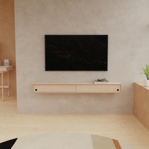 May include: A modern living room featuring a large black television mounted on a light-colored wall. Below the TV is a floating wooden shelf with two drawers. The room has light wood flooring and a neutral color palette.