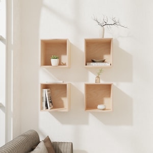 May include: Four light wood square wall shelves are mounted on a white wall. Each shelf displays a decorative item, such as a small plant, books, and a vase with branches. The shelves are arranged in a grid pattern, creating a modern display.