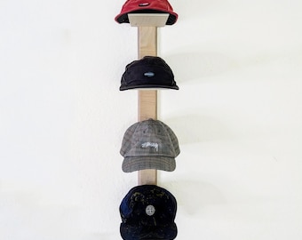 May include: A wooden hat rack displaying four baseball caps. The caps are in various colors, including red, black, gray, and navy blue. The rack is mounted on a white wall, showcasing the hats as a decorative display.