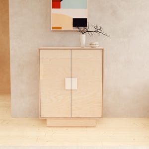 May include: A light wood cabinet with two doors and a square white handle. Above the cabinet is a framed abstract painting with red, black, and blue shapes. A white vase with a branch and a small bowl sit on top of the cabinet.