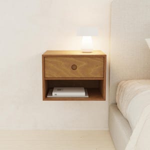 May include: A floating wooden nightstand with a drawer and open shelf. A small white lamp sits on top, illuminating the nightstand. A book rests on the shelf below. The nightstand is mounted on a white wall.
