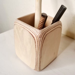 May include: A wooden pencil holder with a square shape and a light wood finish. Two pens are inside the holder.