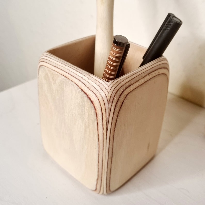 May include: A wooden pencil holder with a square shape and a light wood finish. Two pens are inside the holder.