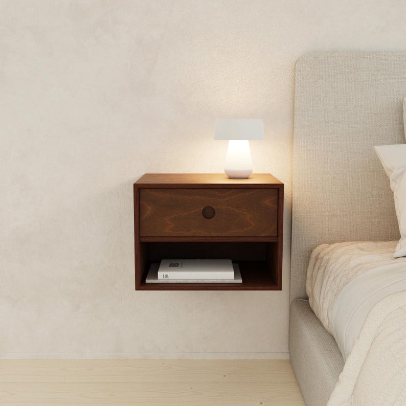 May include: A dark brown floating nightstand with a single drawer and open shelf. A small white lamp sits on top, illuminating the nightstand. A book rests on the shelf below. The nightstand is mounted on a light-colored wall.