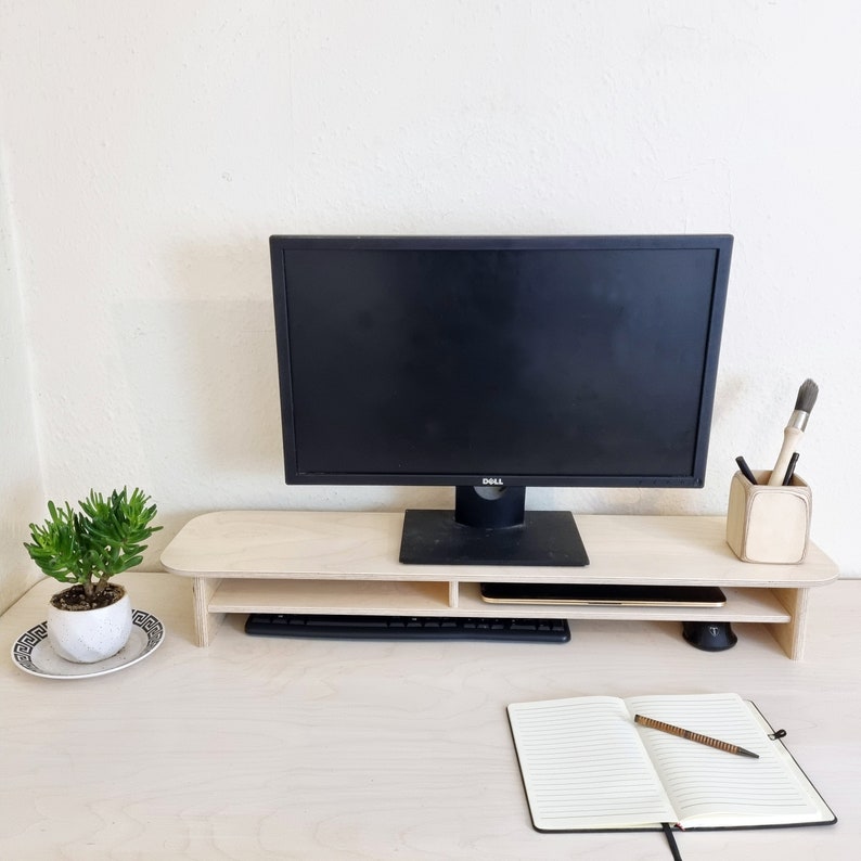 May include: A wooden monitor stand with two shelves, a black computer monitor, a keyboard, a notebook, and a pen. The stand is on a light wood desk.