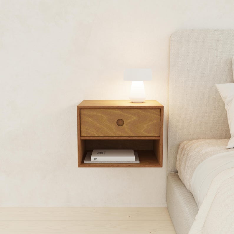 May include: A floating wooden nightstand with a drawer and open shelf. A small white lamp sits on top, illuminating the nightstand. A book rests on the shelf below. The nightstand is mounted on a white wall.