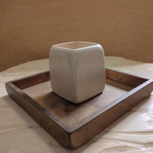 May include: A white wooden square planter with a decorative wood grain pattern sits on a dark brown wooden tray.