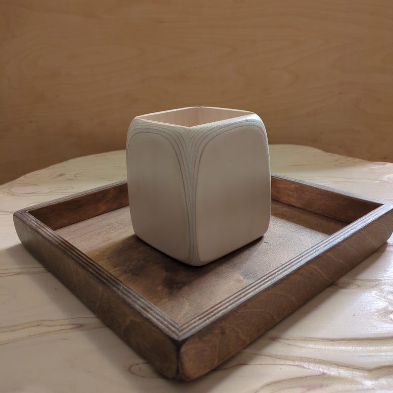 May include: A white wooden square planter with a decorative wood grain pattern sits on a dark brown wooden tray.
