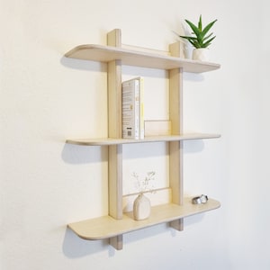 May include: A three-tiered wall shelf made of light wood with rounded edges. The shelf is empty except for a small vase with dried flowers, a watch, and two books titled "Nice is a Big Gift".