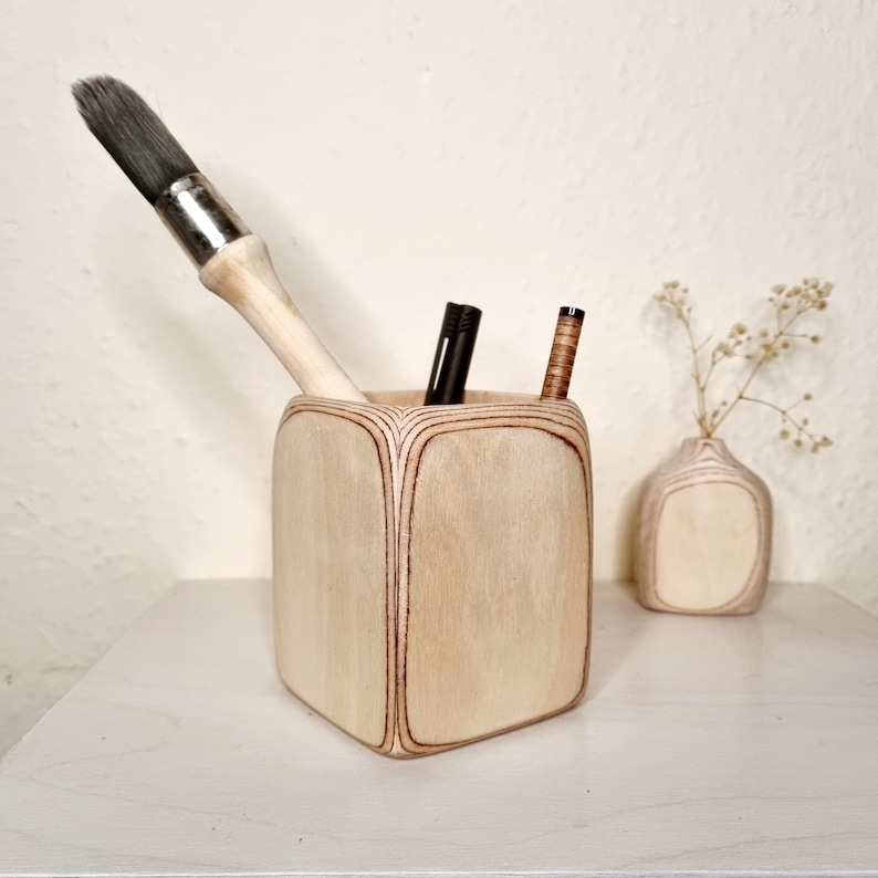 May include: A wooden desk organizer with a square shape and rounded edges. It has a light wood finish and is holding a paintbrush, a black pen, and a brown pen.