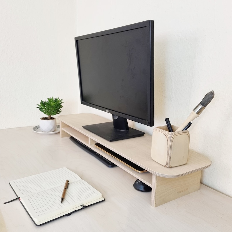 May include: A light wood monitor stand with a black computer monitor, a keyboard, and a pencil holder with a paintbrush and pencils. The stand has a shelf for storage.
