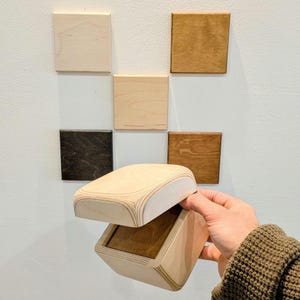 May include: A wooden box with a curved lid is open, revealing a dark interior. The box is light-colored wood with a geometric design. Six square wood samples in various stains are mounted on a white wall.