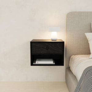 May include: A black floating nightstand with a drawer and open shelf. A small white lamp sits on top, illuminating the surface. A book rests on the shelf below. The nightstand is mounted on a white wall next to a bed.