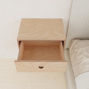 May include: A light-colored wooden floating nightstand with an open drawer. The drawer has a small, oval-shaped handle. The nightstand is mounted on a light-colored wall and sits next to a bed with a light-colored headboard.