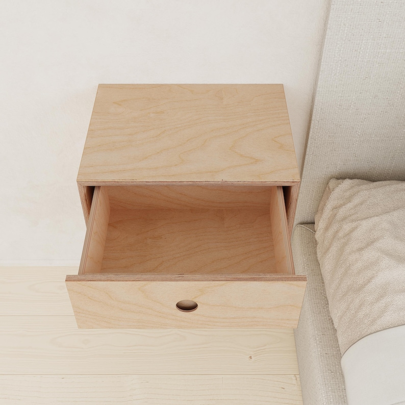 May include: A light-colored wooden floating nightstand with an open drawer. The drawer has a small, oval-shaped handle. The nightstand is mounted on a light-colored wall and sits next to a bed with a light-colored headboard.