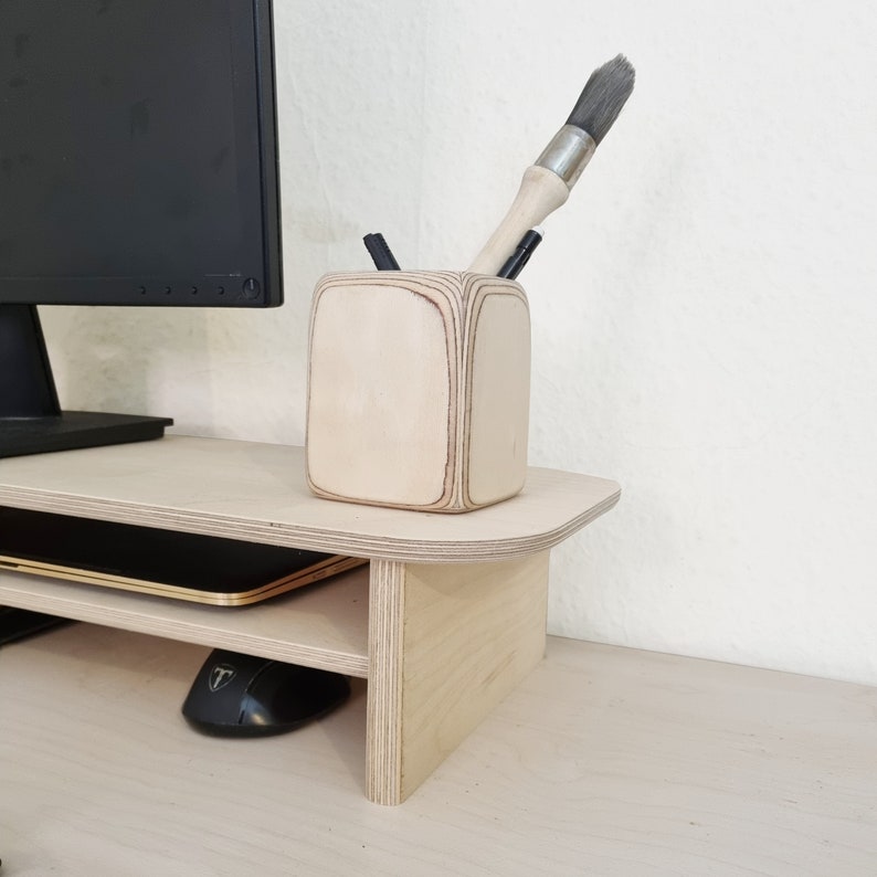 May include: A wooden monitor stand with two shelves and a small wooden pencil holder with a paintbrush and pencils inside. The stand is made of light wood and has a rounded edge.