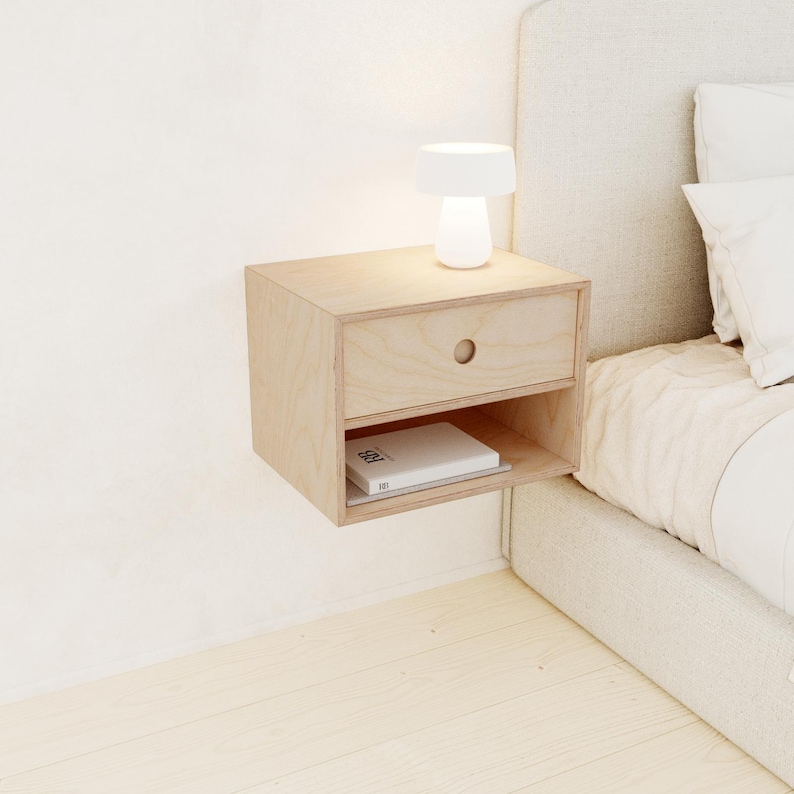 May include: A light-colored, floating nightstand made of wood. It features a drawer with a circular handle and an open shelf below. A small white lamp sits on top, and a book rests on the shelf. The nightstand is mounted on a white wall.