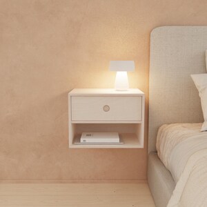 May include: A floating white nightstand with a single drawer and a lower shelf. A small white lamp sits on top, illuminating the nightstand. A book rests on the shelf below. The nightstand is mounted on a peach-colored wall.