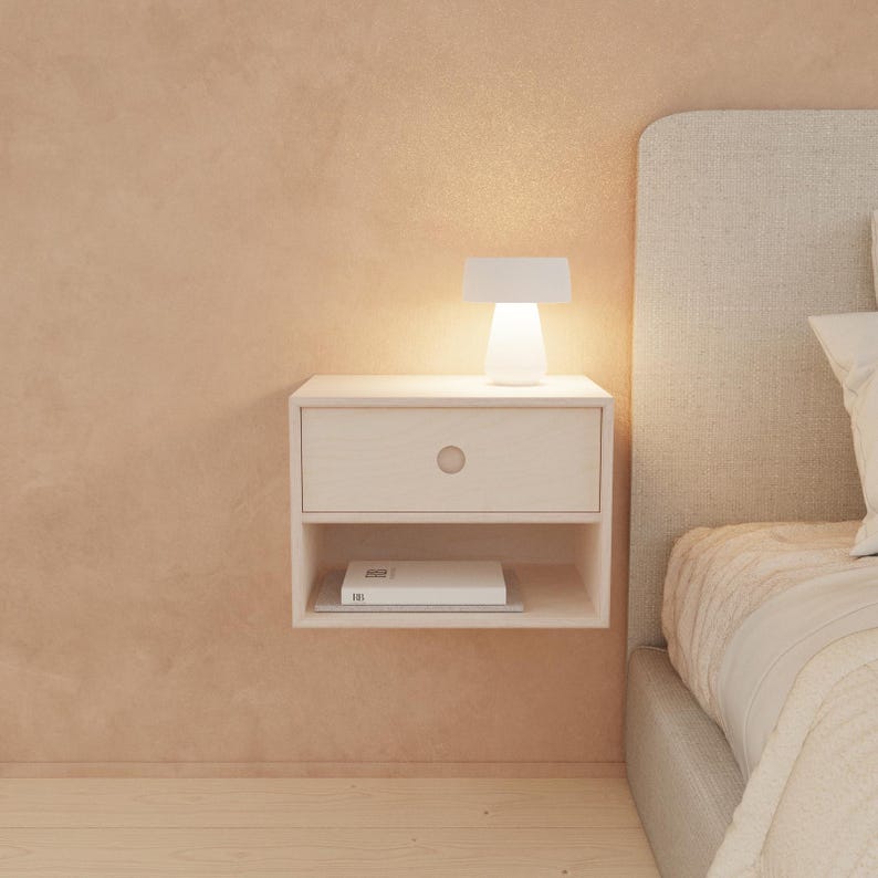 May include: A floating white nightstand with a single drawer and a lower shelf. A small white lamp sits on top, illuminating the nightstand. A book rests on the shelf below. The nightstand is mounted on a peach-colored wall.