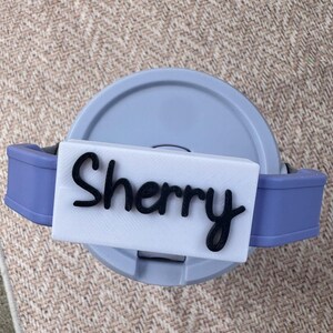 May include: A light blue and gray travel mug with a white rectangular nameplate that reads "Sherry" in black cursive lettering. The mug has a matching light blue handle on each side.