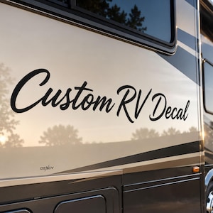 May include: A black vinyl decal with the text Custom RV Decal in a cursive script font applied to the side of a beige and dark gray recreational vehicle. The decal is positioned below a window and above a small logo with the text DEXCV.