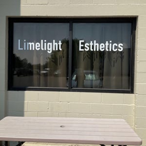 May include: A storefront window with the words "Limelight Esthetics" in white lettering. The window is framed in black and reflects the outside environment. A light brown table is in the foreground, and the building is made of light beige bricks.