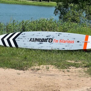 May include: A light blue stand-up paddleboard with black and white chevron stripes on the left side. The board has the word "INFINITY" and the phrase "I'm hilarious" printed on it in black and red.