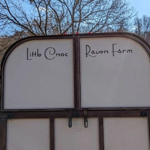 May include: A two-panel sign with the text "Little Cnoc" and "Raven Farm" in black script. The sign has a brown metal frame and white panels. The background includes trees and a blue sky.