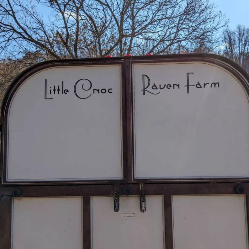 May include: A two-panel sign with the text "Little Cnoc" and "Raven Farm" in black script. The sign has a brown metal frame and white panels. The background includes trees and a blue sky.