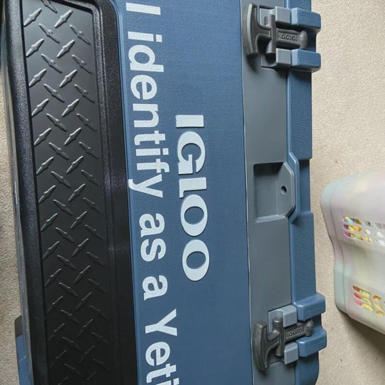 May include: A blue and gray Igloo cooler with the text "I identify as a Yeti" in white. The cooler has a diamond plate pattern on the left side and gray latches. A white and pink object is partially visible on the right.