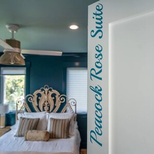 May include: A bedroom with a teal wall and ceiling, a white bed, and a decorative headboard. The door frame on the right has the text "Peacock Rose Suite" in teal script. A ceiling fan is visible.
