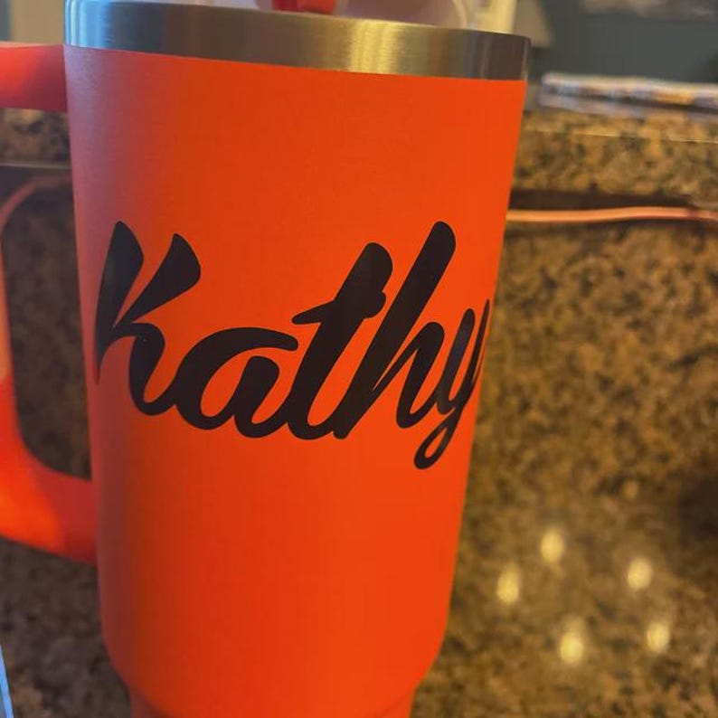 May include: An orange tumbler with a silver rim and a handle. The name "Kathy" is written in black cursive script on the front of the tumbler. The tumbler is likely made of stainless steel.
