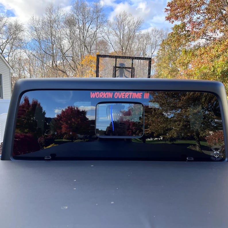 May include: Black truck rear window with the text "WORKIN OVERTIME III" in red. The window reflects a basketball hoop and trees. The truck bed is black.