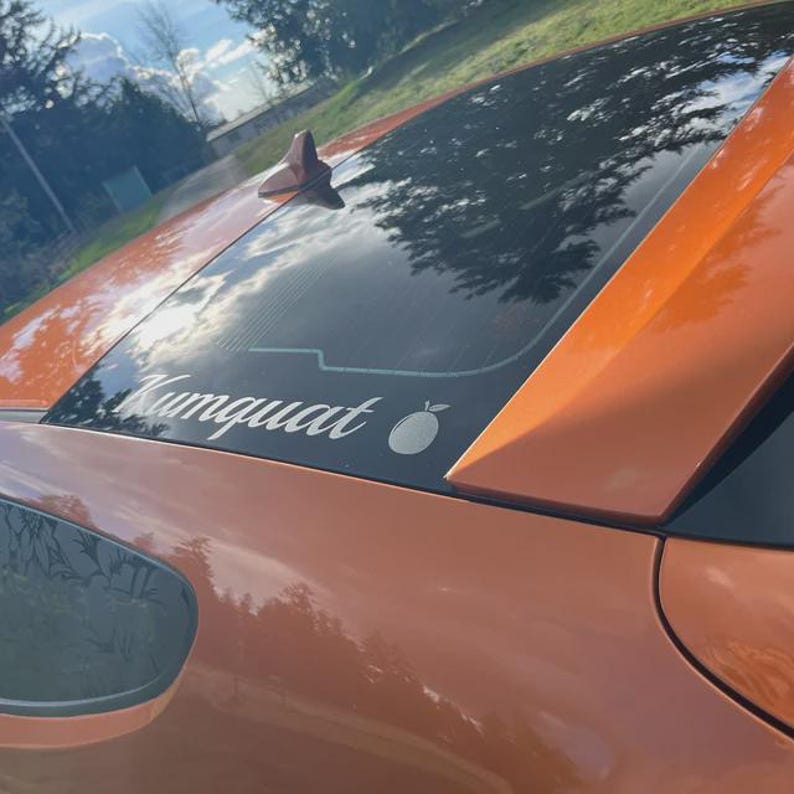 May include: Close-up of an orange car with a black roof. A white decal on the rear window reads "Kumquat" with a small fruit graphic. The car's paint reflects the surrounding environment.