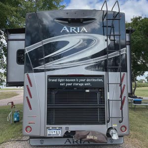 May include: Rear view of a gray and black Aria RV with a ladder and the text "Travel light-heaven is your destination, not your storage unit." The RV has a license plate that reads "319 BZA" and a "Blue Ox" cover.