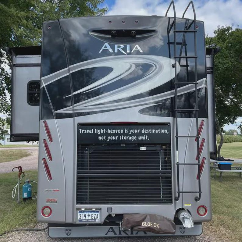 May include: Rear view of a gray and black Aria RV with a ladder and the text "Travel light-heaven is your destination, not your storage unit." The RV has a license plate that reads "319 BZA" and a "Blue Ox" cover.