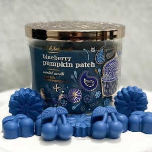 May include: A scented candle in a glass jar with a silver lid, labeled "blueberry pumpkin patch." Surrounding the candle are several blue wax melts in the shapes of cherries, sunflowers, and other floral designs. The candle and melts are on a white surface.