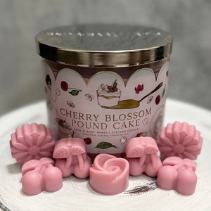 May include: A pink and white candle jar with a silver lid. The jar has a design of a cherry blossom pound cake and the text "Cherry Blossom Pound Cake" and "Bath & Body Works | Scented Candle". The jar is surrounded by six pink wax melts in various shapes, including a cherry, a flower, and a leaf.