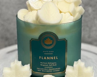 May include: A teal-colored candle with the word "FLANNEL" on the label, surrounded by creamy white wax melts in leaf and other shapes. The candle is from White Barn and is a soy wax blend.