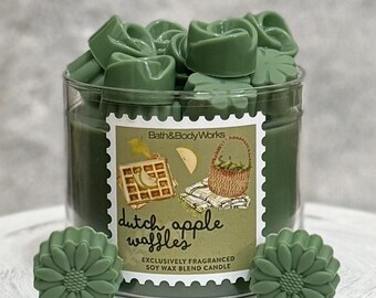 Puede incluir: Una vela de mezcla de cera de soja verde en un tarro de cristal con una etiqueta que dice "Dutch Apple Waffles". La vela está rodeada de ceras perfumadas verdes con forma de flores y hojas. La etiqueta presenta ilustraciones de gofres, manzanas y una cesta.