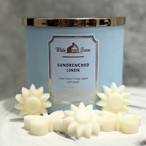 May include: A blue candle jar with a gold lid and a white label that reads "White Barn Sundrenched Linen clean linen - crisp apple soft musk Scented Candle Made with Natural Essential Oils". The candle is surrounded by white wax melts shaped like flowers.