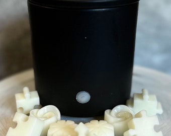 May include: A black electric wax warmer with a white button on the front. The warmer is surrounded by white wax melts shaped like puzzle pieces and flowers.