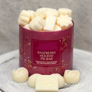 May include: A cylindrical, magenta-colored candle holder filled with white wax melts. The label reads "Raspberry Holiday Pie Bar." Several wax melts are arranged around the base. The candle is a soy wax blend.