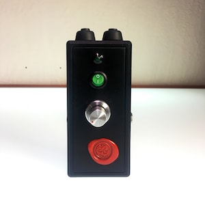 Puede incluir: Caja negra con un sello de cera roja, un botón verde, una perilla plateada y una luz LED verde.