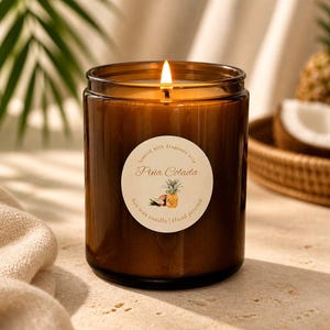 Peut inclure: Une bougie en cire de soja allumée dans un pot en verre ambré cylindrique avec une étiquette ronde blanche. L'étiquette porte le texte Piña Colada, une illustration d'ananas et les mots Scented with fragrance oils et Hand poured.