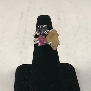 May include: A silver ring with three raw gemstones: a dark blue sapphire, a pink ruby, and a yellow citrine.