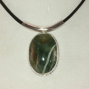 Puede incluir: Un collar de plata con un colgante ovalado de piedra verde y marr&oacute;n. El collar tiene un cord&oacute;n de cuero y una barra de plata.